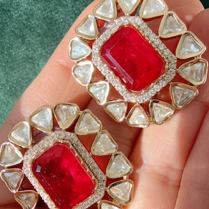 May include: A pair of gold-toned stud earrings. Each earring features a large, rectangular, deep red gemstone surrounded by a halo of small, clear, triangular stones. The earrings are set against a green background.