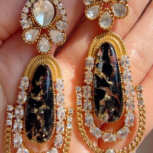 May include: A pair of gold-toned earrings with a black and gold stone center. The earrings feature clear, sparkling crystals and a chain design. The top of the earrings has a cluster of crystals.