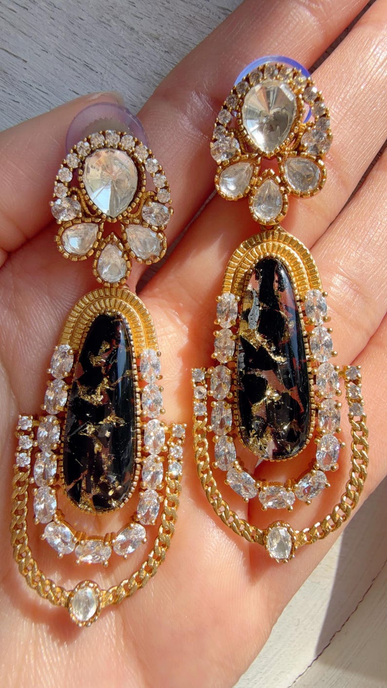 May include: A pair of gold-toned earrings with a black and gold stone center. The earrings feature clear, sparkling crystals and a chain design. The top of the earrings has a cluster of crystals.