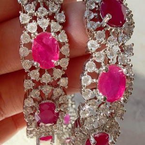 May include: Two silver bangles featuring oval-cut ruby gemstones and small, round, clear gemstones. The rubies are a vibrant pink-red colour, and the smaller stones sparkle. The bangles are ornate and suitable for special occasions.