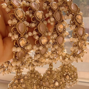 May include: Gold-toned jewelry set featuring ornate earrings and bracelets. The bracelets are adorned with teardrop-shaped, blush-colored stones and small white pearls. The earrings have a similar design, with dangling bell-shaped pendants, creating an elegant, eye-catching look.