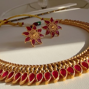 Può includere: Una collana dorata e un set di orecchini abbinati. La collana presenta una serie di accenti rossi a forma di goccia lungo il bordo inferiore. Gli orecchini sono a forma di fiore con accenti rossi.