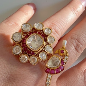 May include: Gold-toned ring with a large, clear, square-cut center stone surrounded by smaller clear stones and red accents. A second ring features a peacock design with clear and red stones.