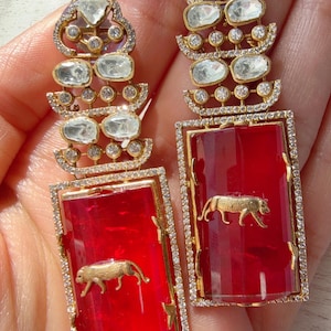 Op de afbeelding: Een paar goudkleurige oorbellen met een rechthoekige rode edelsteen en een gouden tijgerontwerp. De oorbellen hebben een met diamanten bezet frame en een bovenste gedeelte met heldere edelstenen. Een luxe sieraad.