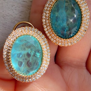 May include: A pair of oval-shaped earrings featuring a large, faceted, translucent turquoise-blue center stone. Each earring is surrounded by a double halo of small, clear, sparkling white stones set in a gold-toned metal frame.