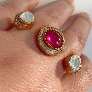 May include: Three gold rings with clear and red gemstones. The rings are set with small clear stones around the larger gemstones. The rings are on a person's hand.