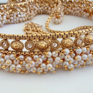 May include: A gold choker necklace with intricate detailing. The necklace is adorned with small, round, white pearls and decorative gold elements. It features circular and oval accents with clear stones.