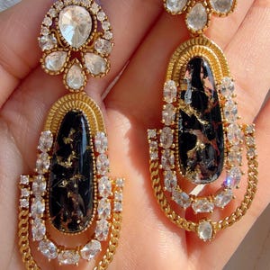 May include: A pair of gold-toned earrings with a black, marbled center stone and clear, sparkling accents. The earrings have a teardrop shape with a chain-like design at the bottom and a floral-shaped top. The earrings are ornate and elegant.