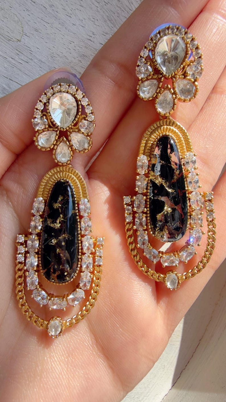 May include: A pair of gold-toned earrings with a black, marbled center stone and clear, sparkling accents. The earrings have a teardrop shape with a chain-like design at the bottom and a floral-shaped top. The earrings are ornate and elegant.