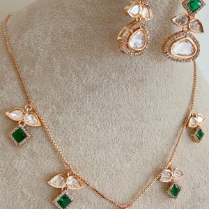 May include: A gold necklace and earrings set with a teardrop-shaped pendant and green stones. The pendant is surrounded by smaller diamonds. The earrings are also teardrop-shaped and have green stones.