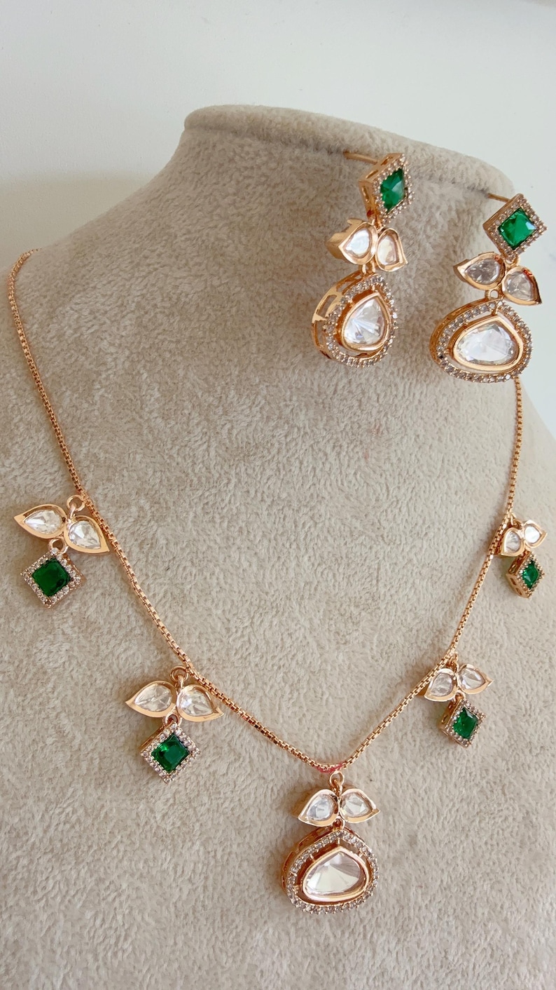 May include: A gold necklace and earrings set with a teardrop-shaped pendant and green stones. The pendant is surrounded by smaller diamonds. The earrings are also teardrop-shaped and have green stones.