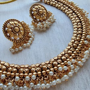 May include: A gold-toned necklace and matching earrings set. The necklace and earrings feature a floral design with dangling faux pearls. The jewelry is displayed on a white surface, showcasing the intricate details of the pieces.
