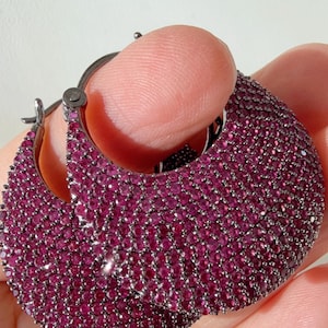 May include: A pair of hoop earrings with a dark metal finish, densely covered in small, round, deep pink gemstones. The earrings are held in a hand, showcasing the intricate detail of the design.