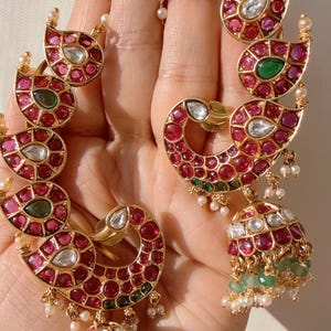 May include: Gold-toned earrings with a peacock design, adorned with red, green, and white gemstones. The earrings feature a jhumka style with dangling green beads and small pearls. The intricate design showcases a blend of traditional and contemporary aesthetics.
