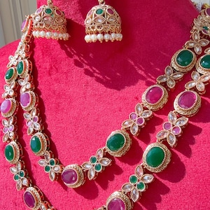 May include: A gold-toned jewelry set featuring a necklace and earrings. The necklace has two strands with alternating green and pink oval gemstones, accented by floral designs. The earrings are bell-shaped with matching gemstones and pearl drops.