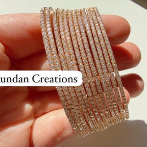 May include: A set of 12 gold bangles, each featuring a row of clear cubic zirconia stones. The bangles are stacked on top of each other and held in a hand. The text "Kundan Creations" is visible in the image.