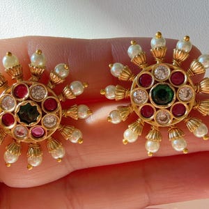 May include: A pair of gold earrings with a circular design featuring a green, red, and white gemstone cluster surrounded by white pearls.
