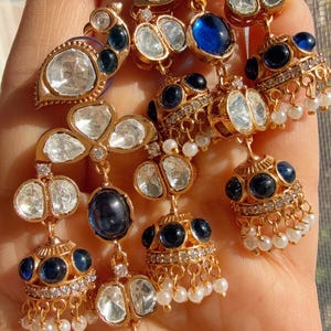 May include: Close-up of gold-toned earrings with clear and dark blue gemstones, and small white pearls. The earrings have intricate designs and are displayed against a soft background. The jewelry is likely for special occasions.