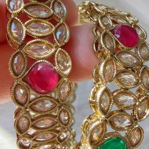 May include: Two gold-toned bangles with intricate designs. Each bangle features oval-shaped clear stones and larger, round red and green gemstones. The jewelry is designed with a floral pattern.