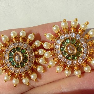 Könnte beinhalten: Ein Paar goldfarbene Ohrringe mit kreisförmigem Design. Jeder Ohrring hat einen zentralen Edelstein, der von kleineren grünen und klaren Steinen und einem Ring aus Perlen umgeben ist. Die Ohrringe haben ein Sonnenstrahl-Design.