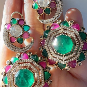 May include: A collection of ornate earrings featuring emerald green, pink, and clear gemstones. The earrings have a gold-tone metal setting and are arranged in floral and geometric designs. The gemstones are surrounded by small, clear crystals.