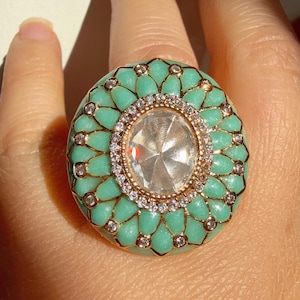 May include: A large, round ring with a turquoise enamel design. The ring features a central, clear, faceted stone surrounded by a ring of small, clear stones. The turquoise enamel is accented with gold-coloured metal.
