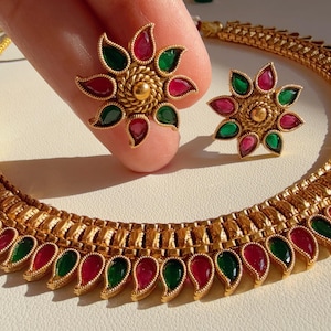 Può includere: Una collana dorata e un set di orecchini abbinati. La collana e gli orecchini presentano un design floreale con accenti rossi e verdi. I gioielli sono esposti su una superficie chiara.