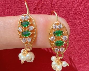 Pode incluir: Brincos dourados com design oval, com pedras preciosas verdes e transparentes. Pequenas pérolas brancas pendem da parte inferior de cada brinco. Fundo vermelho.