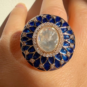May include: An elaborate ring with a central clear gemstone, encircled by smaller clear stones. The gold-toned band is complemented by a deep blue outer ring with a decorative pattern. Jewellery.