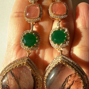 May include: A pair of long, ornate earrings with multiple gemstone settings. The earrings feature turquoise, pink, and green stones, set in gold-colored metal with small, clear crystals. The bottom stones are large and teardrop-shaped.