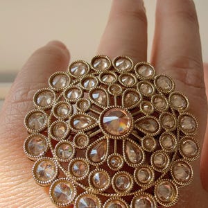 May include: A large, round, gold ring with a circular design of many small, clear stones. The centre stone is larger and has a light brown colour.