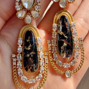 May include: A pair of gold-toned drop earrings. Each earring features a black, teardrop-shaped stone with gold flecks, surrounded by clear, oval-cut crystals. The earrings have a chain-like design and a smaller, round crystal at the top.