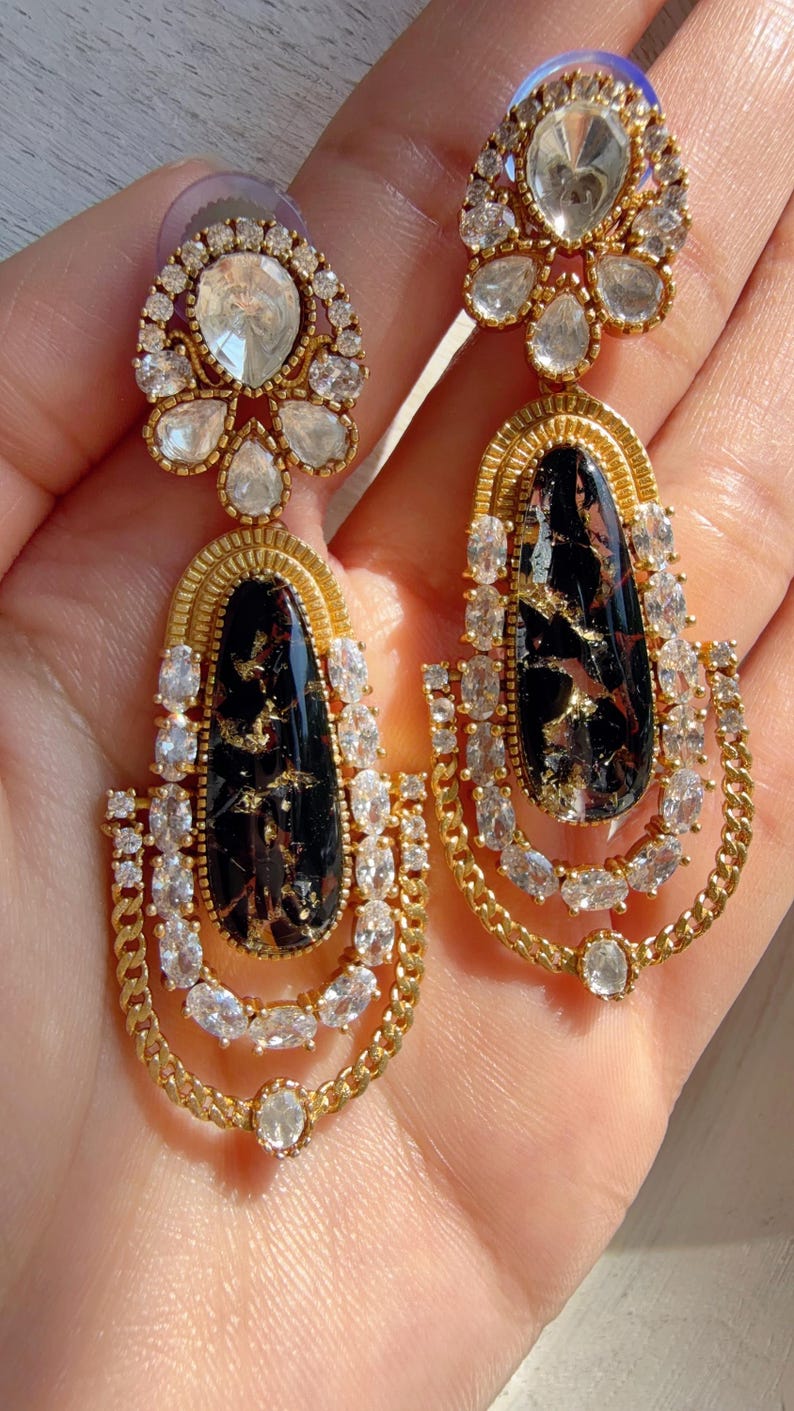 May include: A pair of gold-toned drop earrings. Each earring features a black, teardrop-shaped stone with gold flecks, surrounded by clear, oval-cut crystals. The earrings have a chain-like design and a smaller, round crystal at the top.