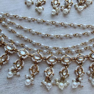 May include: An ornate gold-toned headpiece with multiple strands of small white pearls and decorative pendants. The headpiece features clear, oval-shaped stones and dangling pearl accents, creating an elegant and detailed design. A matching pair of earrings is also visible.