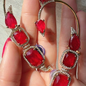 May include: A gold-toned ear cuff featuring a large, curved frame adorned with sparkling clear stones. The design includes multiple vibrant red, faceted, pear-shaped and rectangular gemstones set in gold-toned metal, creating a bold, layered look.