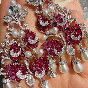 Könnte beinhalten: Elegante silberne Ohrringe mit rubinroten Edelsteinen und weißen Perlen. Die Ohrringe weisen ein florales Design mit roten Steingruppen, funkelnden Akzenten und hängenden Perltropfen auf. Ein luxuriöses Accessoire.