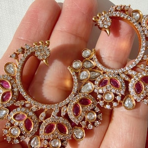 Puede incluir: Pendientes dorados ornamentados con un diseño inspirado en el pavo real. Los pendientes presentan una combinación de piedras preciosas claras y de color rubí, engastadas en un patrón curvo detallado. El diseño incluye un pequeño elemento en forma de pájaro.