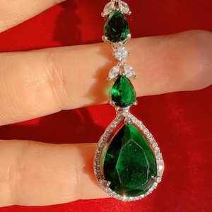 Może przedstawiać: Srebrny naszyjnik z wisiorkiem z serią zielonych kamieni szlachetnych w kształcie łezki. Największy kamień znajduje się na dole, otoczony aureolą małych, przezroczystych kryształków. Naszyjnik jest umieszczony na czerwonym tle.