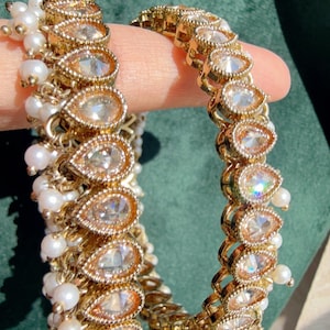 May include: Two gold-toned bangles with teardrop-shaped clear stones and small white pearls. The bangles have a textured gold frame around the stones. The pearls are clustered and dangle from the bangles. Jewelry.