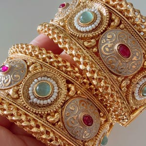May include: Three gold bangles with intricate designs and colourful gemstones. The bangles are stacked on top of each other and feature a mix of red, green and white stones.