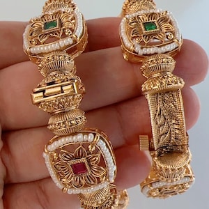 Könnte beinhalten: Ein Paar goldene Armreifen mit aufwendigen Designs und Perlenakzenten. Ein Armreif hat einen grünen Stein, während der andere einen roten Stein hat.