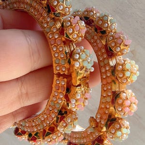 May include: A pair of gold-toned hinged bangles featuring intricate metalwork, inlaid with small white, pink, and light green beads. The design includes triangular accents in red, green, and black enamel, with a textured surface throughout the metal.