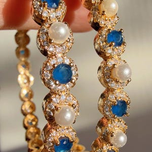 May include: Gold-toned hoop earrings featuring alternating blue gemstones and white pearls, each surrounded by small, clear crystals. The earrings have a detailed, ornate design, with a warm, reflective finish.