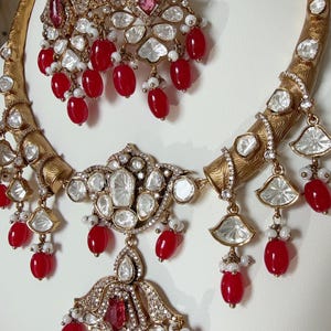 May include: A gold necklace and earrings set with red and white beads, clear stones, and a pink stone. The necklace has a curved design and the earrings are dangle style.