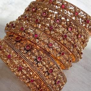 May include: Gold-toned bangles with intricate floral designs and small red gemstone accents. The bangles are stacked, showcasing the detailed craftsmanship and ornate style. These accessories are suitable for special occasions.