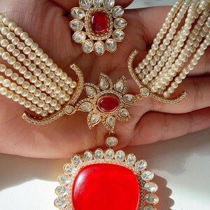 Op de afbeelding: Een goudkleurige sieradenset met een grote rode edelsteen hanger omringd door heldere kristallen. De set bevat een ring en een ketting met meerdere strengen parels. Het ontwerp bevat bloemen en geometrische elementen.