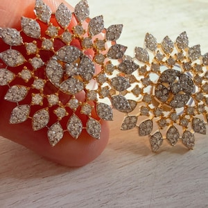 May include: Two gold-toned, floral-shaped earrings. Each earring features a central cluster of small, round diamonds surrounded by leaf-shaped diamond accents. The earrings are held against a light-colored surface.