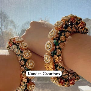 May include: Two gold bangles with green, red, and white enamel and pearl accents. The bangles are decorated with round, raised, jeweled stones. The text "Kundan Creations" is visible in the image.