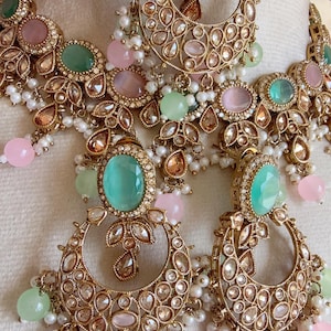 Könnte beinhalten: Ein aufwendiges, goldfarbenes Schmuckset mit Halskette und Ohrringen. Das Set ist mit großen, ovalen tealfarbenen Edelsteinen, kleineren rosa und grünen Steinen und baumelnden Perlenakzenten verziert. Die Ohrringe haben eine Halbmondform.
