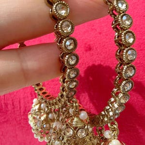 May include: Gold-toned, ornate bangles with clear, round gemstones and dangling bell-shaped charms. The charms are adorned with small pearls. The jewelry is displayed against a bright pink background.